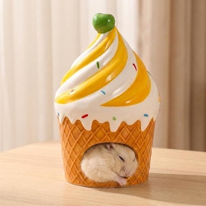 May include: A ceramic hamster house shaped like an ice cream cone. The cone is a waffle pattern, and the ice cream has yellow and white swirls with colorful sprinkles. A small, white hamster is sleeping inside the house.