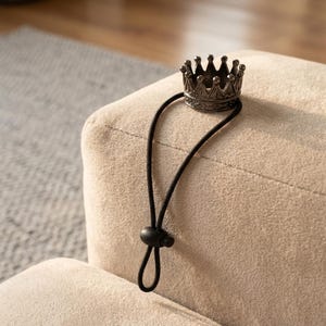 May include: A small, antique-style crown hair tie in a dark metallic finish. The crown is attached to a black elastic band with a small, round toggle. The hair accessory is resting on a beige, textured surface.