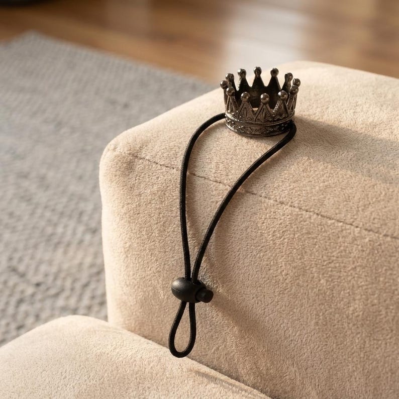 May include: A small, antique-style crown hair tie in a dark metallic finish. The crown is attached to a black elastic band with a small, round toggle. The hair accessory is resting on a beige, textured surface.