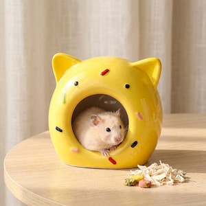 May include: A yellow ceramic hamster house shaped like a donut with cat ears. The house has black and red sprinkles and a small hamster inside. A pile of food and bedding sits on the table.