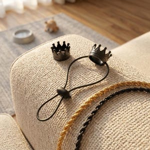 May include: Two black crown-shaped wine bottle stoppers. One stopper is attached to a black cord. A gold and black rope are in the foreground. The items are on a beige upholstered surface.