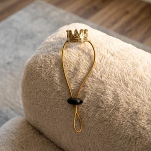 May include: A gold-colored crown-shaped accessory with a loop and a black bead. The crown is detailed with small points and a textured surface. The accessory is attached to a soft, beige, plush surface.