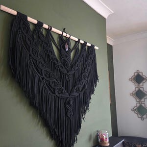 Puede incluir: Un gran tapiz de macramé negro con una barra de madera. El intrincado diseño presenta patrones anudados y largas franjas, creando una estética texturizada y bohemia. Un pequeño colgante de cristal cuelga de la barra.