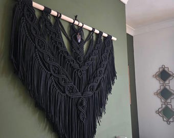 May include: A large, black macrame wall hanging with a wooden dowel. The intricate design features knotted patterns and long fringe, creating a textured, bohemian aesthetic. A small crystal pendant hangs from the dowel, adding a touch of sparkle.