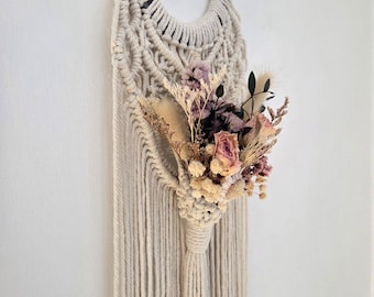 May include: A wall hanging featuring a dark gray metal horseshoe wrapped in off-white macrame cord with intricate knotting. A bouquet of dried flowers in shades of pink, purple, and cream is tucked into the center, with long fringe hanging below.