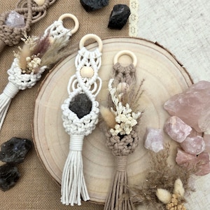 Pode incluir: Pingentes de carro em macramé com anéis de madeira, com cristais e flores secas. Os pingentes são em tons de branco, bege e castanho, com uma estética natural. Cristais pretos e rosa também estão presentes.