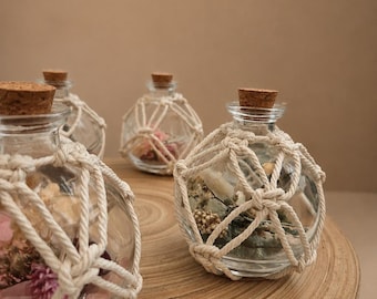 May include: Three clear glass spherical bottles with cork stoppers, each encased in a decorative off-white macrame cord netting. Inside each bottle are various dried flowers and botanical elements in shades of pink, green, and cream.