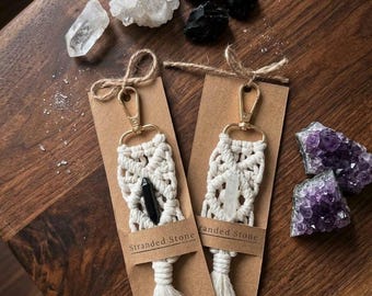 May include: Two macrame keychains with gold-tone lobster clasps, each featuring a central crystal point and a bottom tassel. The keychains are attached to brown cardboard tags labeled Stranded Stone Designs, surrounded by clear, black, and purple crystals.