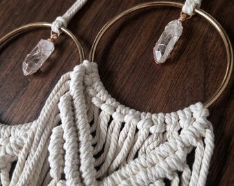 May include: A macrame wall hanging featuring off-white cotton cord knotted into a geometric pattern. Two large gold-toned metal rings are integrated into the design, each suspending a clear, raw crystal point wrapped in thin gold-colored wire.