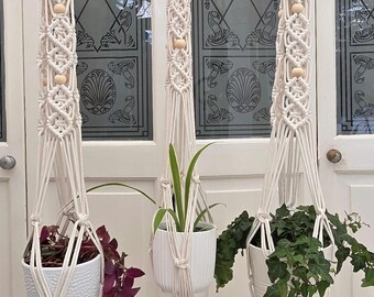 May include: Three macrame plant hangers with wooden rings and beads. Each off-white hanger holds a white ceramic pot with greenery. The plants include a trailing purple plant, a green plant with long leaves, and a green ivy plant.