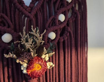 May include: A deep burgundy macrame wall hanging featuring intricate knotted patterns, smooth white wooden beads, and a central arrangement of dried flowers including a vibrant orange strawflower, sprigs of greenery, and small white blossoms.