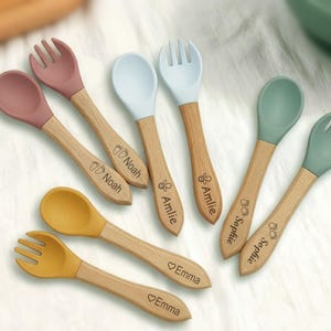 May include: A collection of baby spoons and forks with wooden handles and colorful silicone heads. The utensils are in shades of pink, blue, green, and yellow. Each handle is engraved with a name. The spoons and forks are designed for infant feeding.