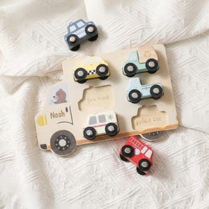May include: A wooden puzzle board shaped like a large truck with the name Noah printed on it. It includes several small wooden vehicle toys: a blue car, a yellow taxi, a green truck, a light blue truck, an ambulance, and a red fire truck.