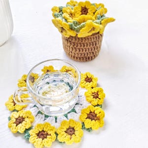 Puede incluir: Un posavasos de ganchillo con diseño de girasol, centro blanco y pétalos amarillos, que sostiene una taza de cristal transparente con agua, junto a una pequeña maceta de ganchillo marrón llena de girasoles amarillos y verdes.