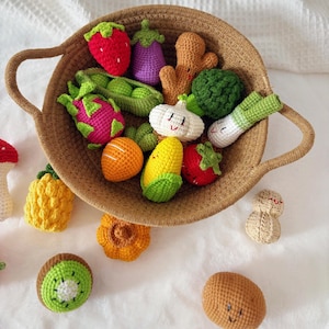 Puede incluir: Una colección de frutas y verduras de ganchillo con caras bordadas, incluyendo fresa, berenjena, jengibre, brócoli, ajo, puerro, fruta del dragón, guisantes, maíz y naranja, dentro de una cesta redonda de cuerda trenzada color canela.