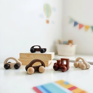May include: A collection of five small wooden toy vehicles, including four cars with rounded arches and one train engine. The toys are crafted from light and dark wood, featuring smooth surfaces and circular wheels, arranged on a white surface.