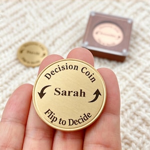 May include: A circular gold-toned metal decision coin featuring the engraved text "Decision Coin" at the top, "Sarah" in the centre, and "Flip to Decide" at the bottom, accompanied by two curved arrows pointing towards the centre name.