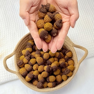 Op de afbeelding: Een verzameling kleine, gehaakte eikels met goudbruine noten en donkerbruine dopjes. De eikels worden gepresenteerd in een ronde, geweven beige mand met twee handvatten, waarbij nog enkele eikels in open handpalmen worden vastgehouden.