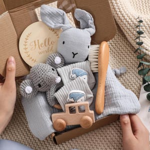 Puede incluir: Un juego de regalo para bebé en una caja de cartón, que incluye un peluche de conejo gris, un elefante de ganchillo, calcetines con un diseño de oso durmiente, un coche de madera, un cepillo de madera y un letrero de madera que dice "Hello world". El juego está envuelto en una tela de muselina azul claro.