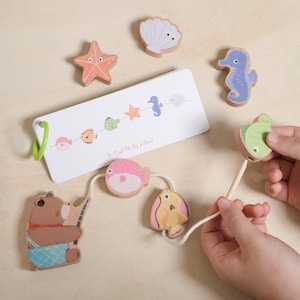 Op de afbeelding: Houten speelgoedset met zeedieren, waaronder een zeester, zeepaardje en vissen. Een berenfiguur houdt een hengel vast en een kaart toont illustraties van zeedieren. De set is ontworpen voor kinderen.