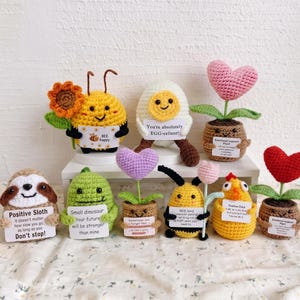 May include: A collection of nine small crocheted figures including a bee with a sunflower, an egg, a heart-shaped plant, a sloth, a dinosaur, a purple heart plant, a bee with a flower, a chick, and a red heart plant, each holding a white sign.