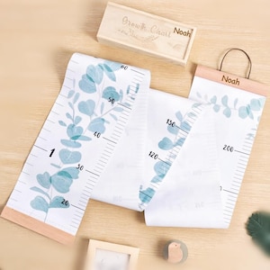 May include: A white canvas growth chart with a blue-green eucalyptus leaf pattern, black measurement markings, and wooden dowels. The chart features the name Noah and is accompanied by a matching wooden storage box labelled Growth Chart Noah.