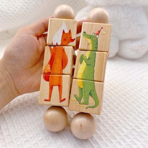 May include: Two vertical wooden toy columns, each consisting of three stacked square blocks and two rounded end pieces. One column features an orange fox illustration, while the other displays a green crocodile wearing a small red party hat.