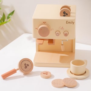 May include: A wooden toy coffee maker set in a light beige color. The set includes a coffee machine with the name "Emily" and the word "Coffee" printed on it, a portafilter, a cup, a saucer, and two cookies. The coffee machine has a wooden handle and a dial.