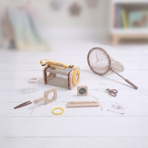 May include: A wooden bug catching kit with a yellow strap, a net, tweezers, a magnifying glass, and a small compass. The kit includes a small container, a ruler, and a yellow rubber band. The items are arranged on a white wooden surface.
