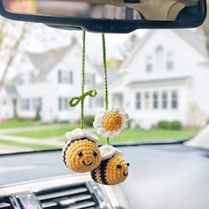 Op de afbeelding: Een hangend auto-accessoire met twee gehaakte bijen met geel-zwart gestreepte lichamen, witte vleugels en zwarte kraaloogjes, opgehangen aan groene garen koorden naast een witte gehaakte bloem met een geel hart.