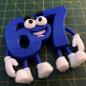 May include: A blue number 67 figurine with a cartoon face, featuring large white eyes with black pupils, black eyebrows, a wide smiling mouth with white teeth, and a small pink tongue. The number has blue arms and legs with white gloves and shoes.
