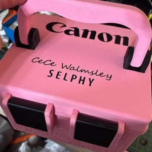 Puede incluir: Un maletín de transporte rectangular de color rosa claro con un asa superior y dos cierres frontales negros. La superficie superior presenta el texto negro Canon, Cece Walmsley y SELPHY. El estuche tiene un acabado texturizado y bisagras negras.