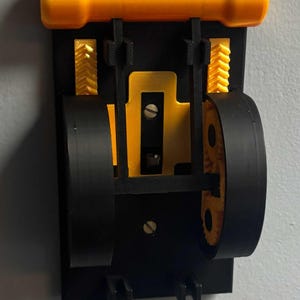 May include: A wall-mounted mechanical device featuring a black rectangular base with a horizontal yellow cylindrical top bar. The assembly includes two large black wheels, yellow gear components, and a central yellow plate secured by two silver screws.