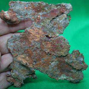 May include: A piece of raw copper ore with a rough, irregular shape. The surface displays a mix of colors, including reddish-brown, gray, and hints of green. The mineral is held in a hand against a green background.