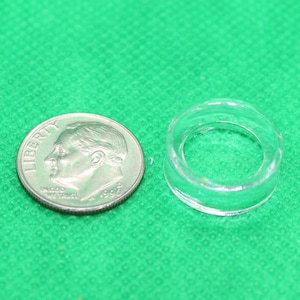 May include: A clear, circular plastic object next to a U.S. dime coin on a green surface. The plastic object appears to be a small ring or washer. The dime shows the profile of Franklin D. Roosevelt.