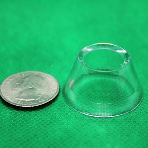 May include: A clear, conical glass object with a circular opening at the top, next to a US quarter coin. The object is transparent, and the coin is silver. The background is a solid green fabric.