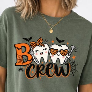 Könnte beinhalten: Ein olivgrünes T-Shirt mit einem Grafikdruck und dem Text "BOO CREW". Das Design zeigt zwei Cartoon-Zähne, einer mit einer Leopardenmuster-Schleife und der andere mit herzförmiger Leopardenmuster-Sonnenbrille, umgeben von Fledermäusen und Spinnennetzen.