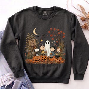 K&ouml;nnte beinhalten: Ein anthrazitfarbenes Sweatshirt mit Rundhalsausschnitt und einer Grafik von Peanuts-Figuren, die auf K&uuml;rbissen sitzen. Das Design zeigt eine Mondsichel, ein Schild mit der Aufschrift PUMPKIN SPICE & AUTUMN ADVENTURES, ein Gespenst und Herbstlaub.