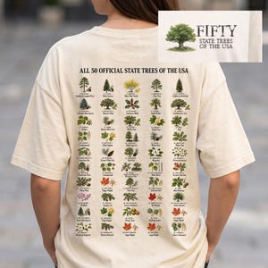 May include: A cream-colored t-shirt featuring a detailed graphic on the back titled ALL 50 OFFICIAL STATE TREES OF THE USA. The design displays fifty small, labeled illustrations of various trees and foliage, with a logo at the top showing a tree.