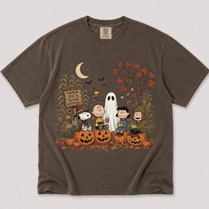 K&ouml;nnte beinhalten: Ein dunkelbraunes Kurzarm-T-Shirt mit einer Grafik von Peanuts-Figuren, die auf K&uuml;rbissen sitzen. Der Aufdruck zeigt ein Schild mit der Aufschrift PUMPKIN SPICE & AUTUMN ADVENTURES, eine Mondsichel, Flederm&auml;use, ein Gespenst und Herbstlaub.
