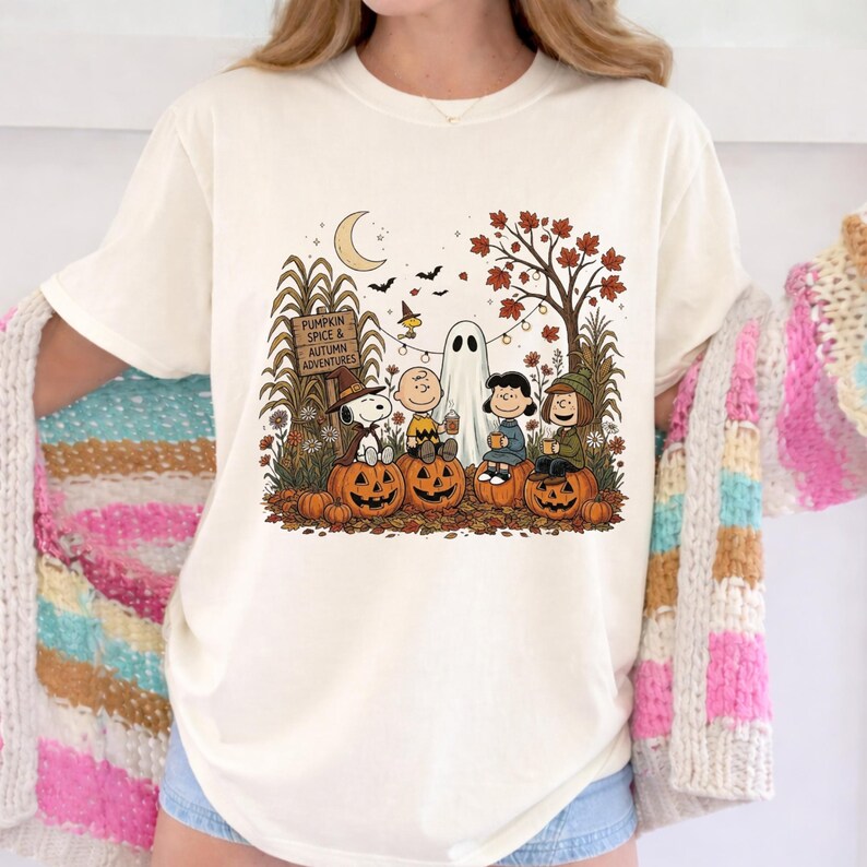 K&ouml;nnte beinhalten: Ein cremefarbenes T-Shirt mit einer Grafik von Peanuts-Figuren, die auf K&uuml;rbissen in einer herbstlichen Szene sitzen. Das Design zeigt ein Schild mit der Aufschrift PUMPKIN SPICE & AUTUMN ADVENTURES, ein Gespenst, Flederm&auml;use und Herbstlaub.