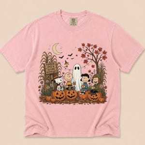 K&ouml;nnte beinhalten: Ein hellrosa T-Shirt mit einer Grafik von Peanuts-Figuren, die in einer herbstlichen Umgebung auf K&uuml;rbissen sitzen. Das Design zeigt ein Schild mit der Aufschrift PUMPKIN SPICE & AUTUMN ADVENTURES, einen Geist, eine Mondsichel und Laub.