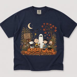 K&ouml;nnte beinhalten: Ein marineblaues T-Shirt mit einer Grafik von Peanuts-Figuren, die auf K&uuml;rbissen in einem herbstlichen Feld sitzen. Das Design zeigt ein Holzschild mit der Aufschrift PUMPKIN SPICE & AUTUMN ADVENTURES, eine Mondsichel, ein Gespenst und Herbstlaub.