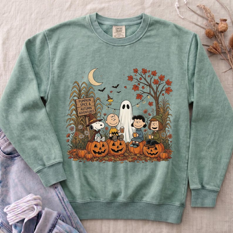K&ouml;nnte beinhalten: Ein salbeigr&uuml;nes Sweatshirt mit Rundhalsausschnitt und einem Grafikdruck der Peanuts-Figuren, die Halloween feiern. Das Design zeigt Snoopy, Charlie Brown, Lucy und Linus, die auf K&uuml;rbissen unter einer Mondsichel und einem Herbstbaum sitzen.