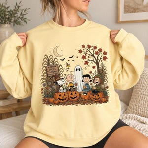 K&ouml;nnte beinhalten: Ein blassgelbes Sweatshirt mit Rundhalsausschnitt und einem Grafikdruck von Peanuts-Figuren, die in einer herbstlichen Umgebung auf K&uuml;rbissen sitzen. Der Text lautet PUMPKIN SPICE & AUTUMN ADVENTURES. Die Szene zeigt ein Gespenst, einen Baum und Flederm&auml;use.