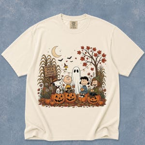 K&ouml;nnte beinhalten: Ein cremefarbenes T-Shirt mit einer Grafik von Peanuts-Figuren, die in einer herbstlichen Szene auf K&uuml;rbissen sitzen. Das Design zeigt ein Schild mit der Aufschrift PUMPKIN SPICE & AUTUMN ADVENTURES, ein Gespenst, eine Mondsichel und Herbstlaub.