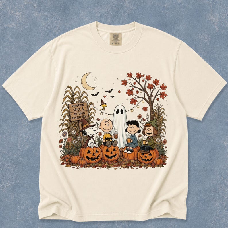 K&ouml;nnte beinhalten: Ein cremefarbenes T-Shirt mit einer Grafik von Peanuts-Figuren, die in einer herbstlichen Szene auf K&uuml;rbissen sitzen. Das Design zeigt ein Schild mit der Aufschrift PUMPKIN SPICE & AUTUMN ADVENTURES, ein Gespenst, eine Mondsichel und Herbstlaub.