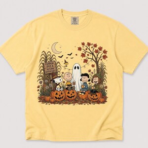 K&ouml;nnte beinhalten: Ein blassgelbes T-Shirt mit einer Grafik von Peanuts-Figuren, die auf K&uuml;rbissen in einer herbstlichen Szene sitzen. Das Design zeigt ein Schild mit der Aufschrift PUMPKIN SPICE & AUTUMN ADVENTURES, einen Geist, einen Mond und Herbstlaub.