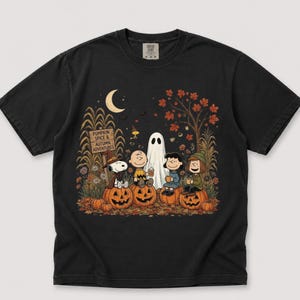 K&ouml;nnte beinhalten: Ein schwarzes Kurzarm-T-Shirt mit einer Grafik der Peanuts-Figuren in einem K&uuml;rbisfeld. Das Design zeigt Snoopy, Charlie Brown, Lucy und Linus mit einem Geist, K&uuml;rbissen und einem Schild mit der Aufschrift PUMPKIN SPICE & AUTUMN ADVENTURES.