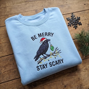 May include: A light blue crewneck sweatshirt featuring an embroidered black crow wearing a red Santa hat, holding a green Christmas light bulb in its beak while perched on a branch wrapped in colorful string lights, with the text BE MERRY STAY SCARY.